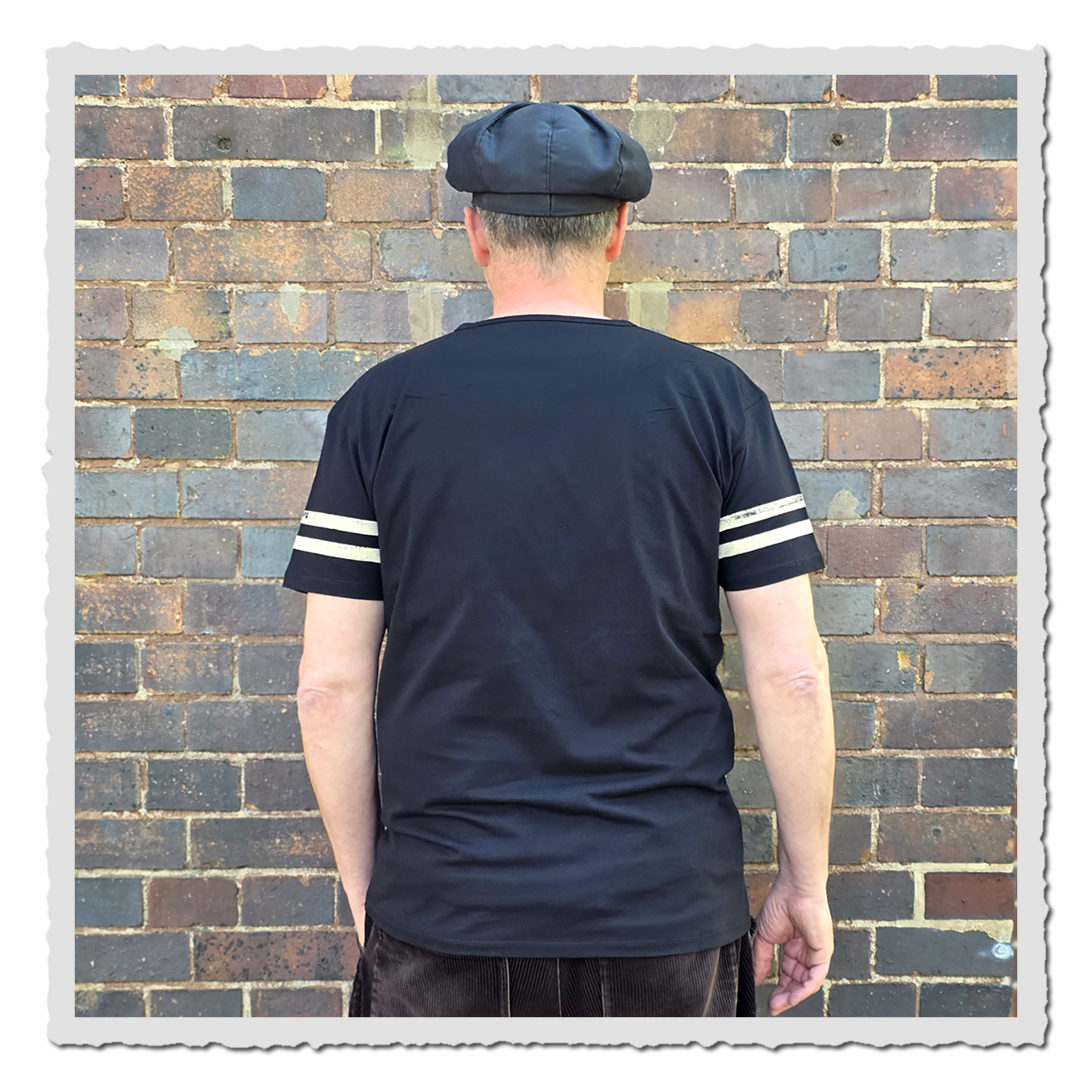 Person von hinten mit schwarzem Kurzarmshirt und Ärmelstreifen vor Backsteinwand.